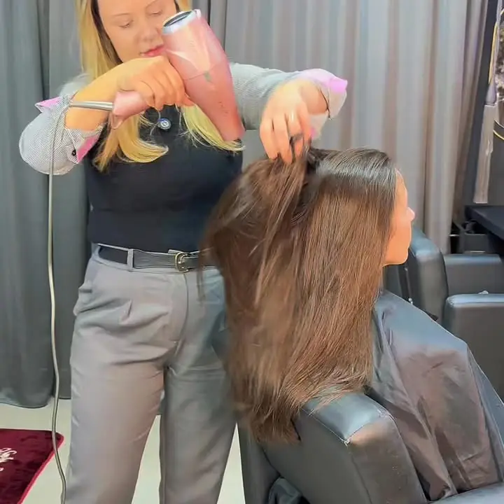 A Fabi com secador rosa e escova trabalhando o cabelo castanho de uma cliente sentada no salão